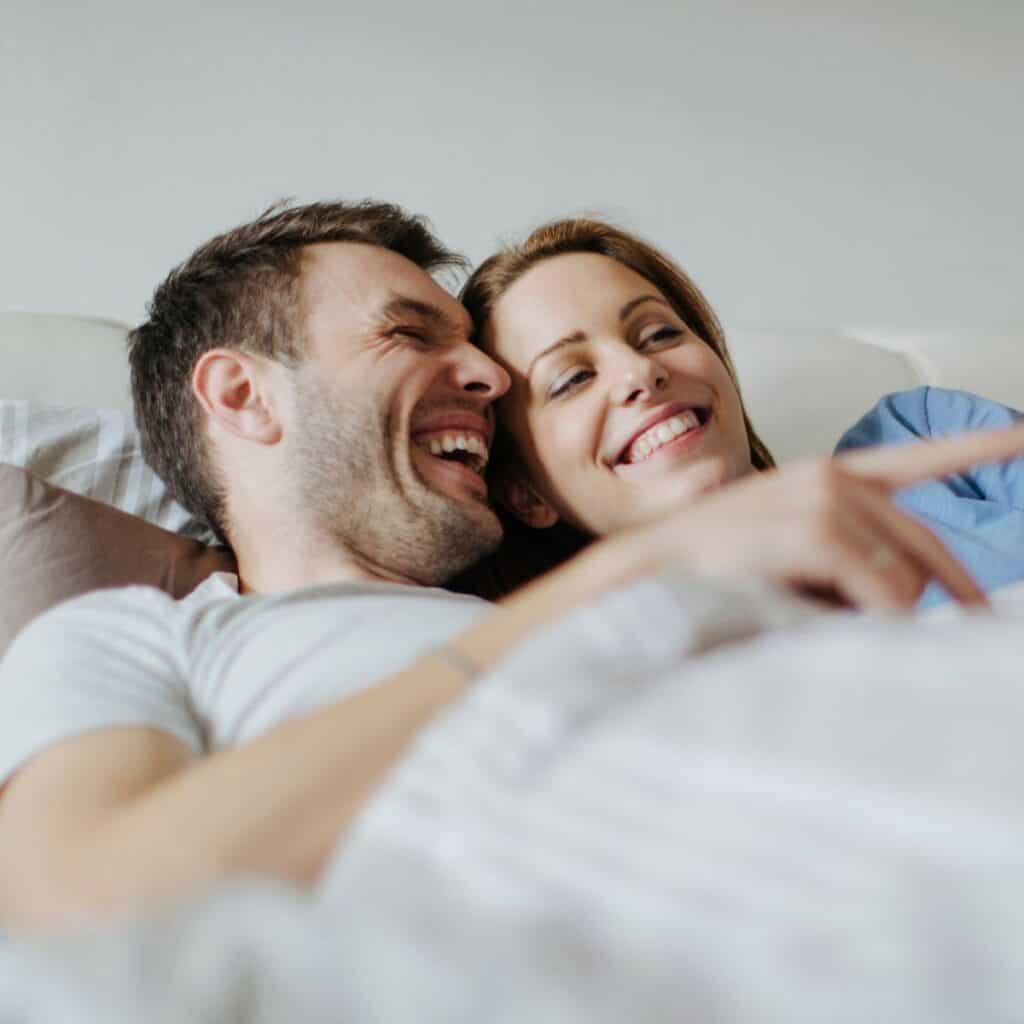 A couple cuddling and laughing on the bed, asking each other these dirty never have i ever questions for couples