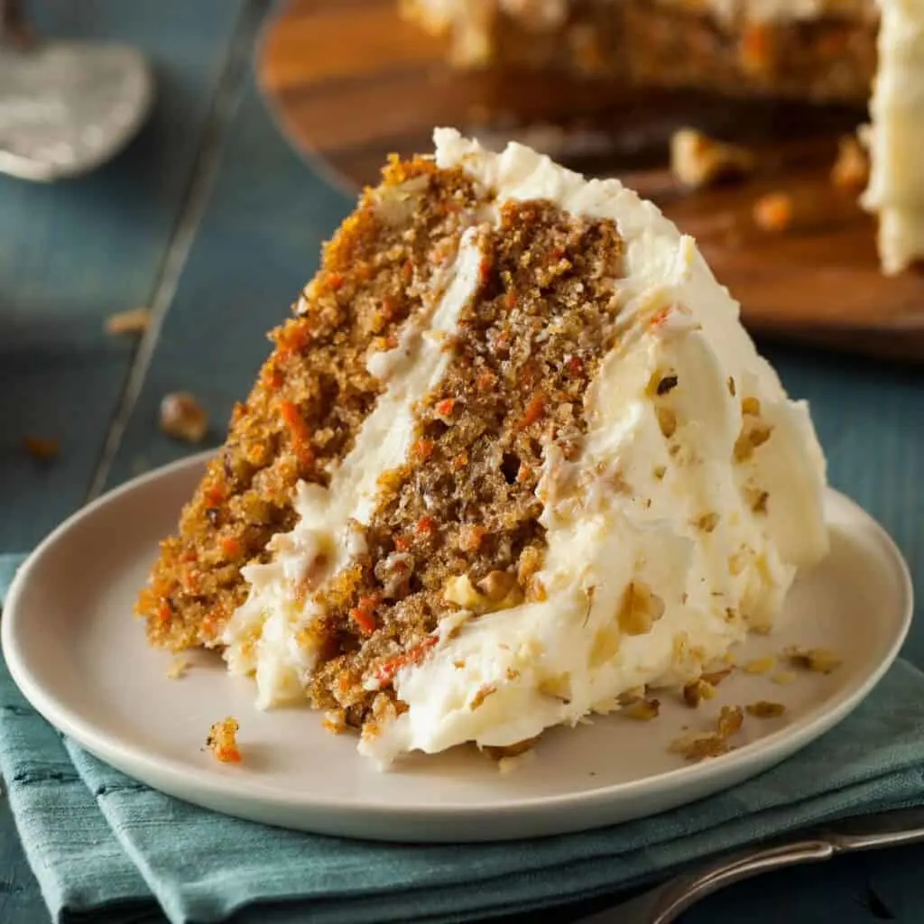 A slice of carrot cake on a plate next to a carrot cake.