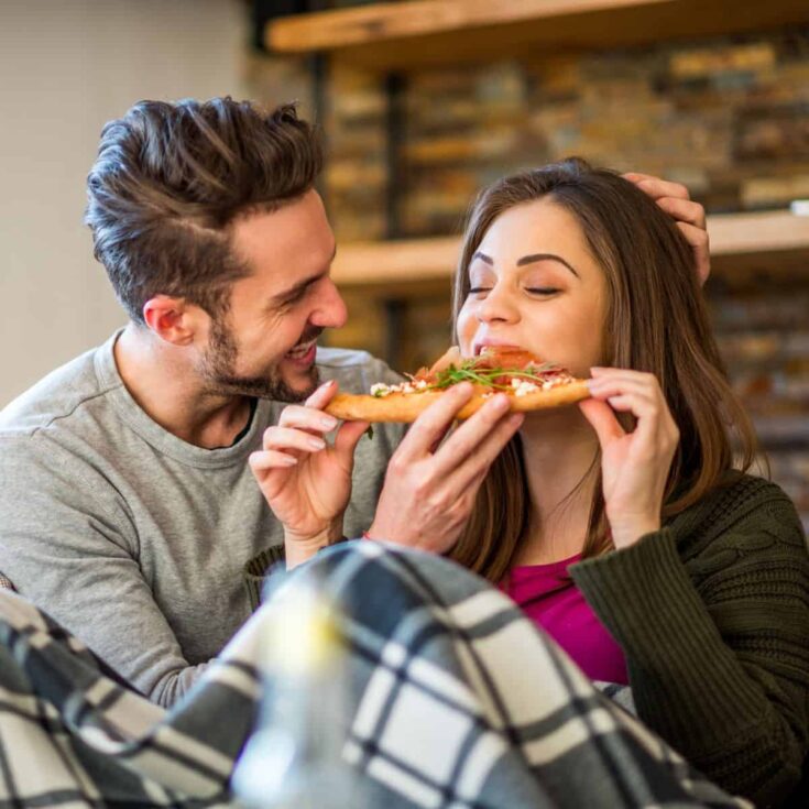 A happy couple having a DIY pizza date night, the man feeding the woman while they are sitting on the couch