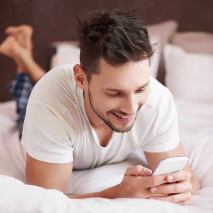 A man lying on the bed, smiling while looking at his phone, texting some good morning love messages