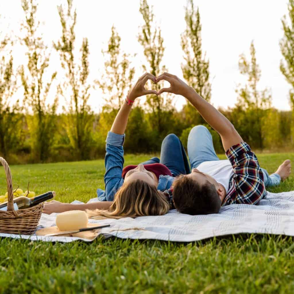 A couple lying on their backs on a picnic blanket making a love heart shape with their fingers.