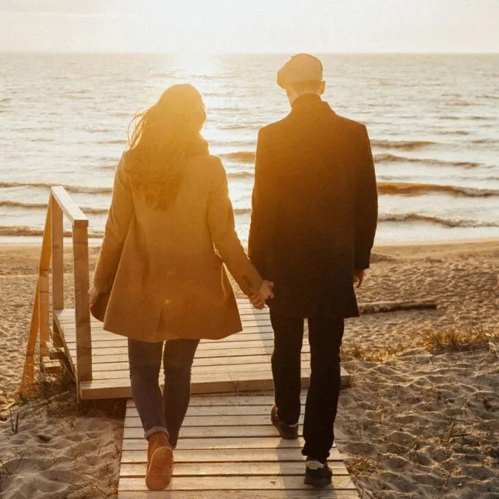 A couple walking hand in hand toward the sea at sunset asking each other relationship questions to keep connected.