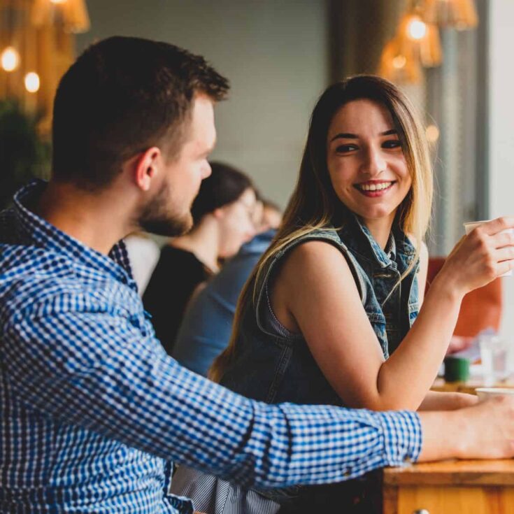 A couple talking to each other in a caf&eacute;, the woman happily looking at the man while holding a coffee, both asking each other these get to know you questions