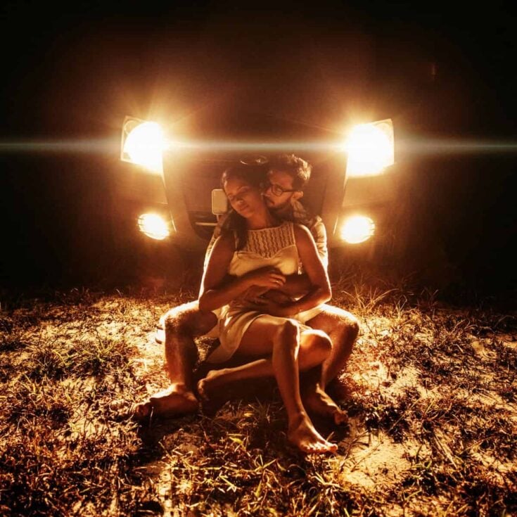 Loved up couple doing one of these late night date ideas: she is sitting cradled by him by the lights of their car