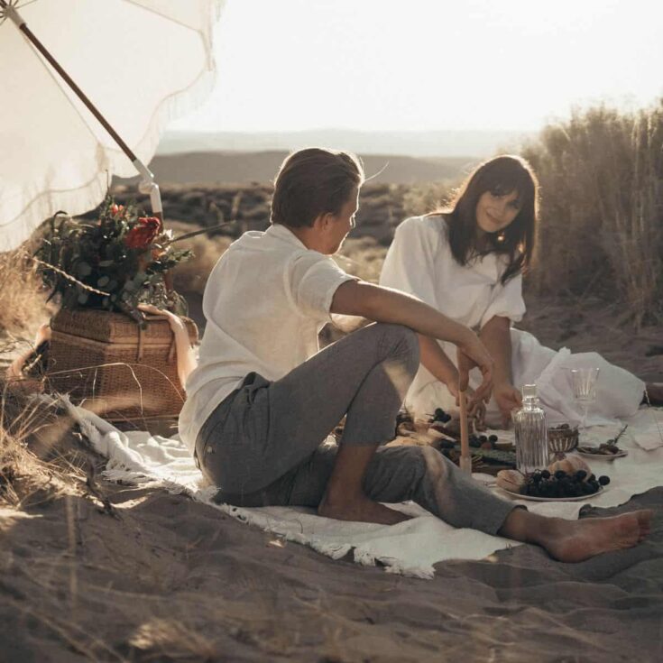 Couple under an umbrella having a picnic at the beach: picnic date ideas.