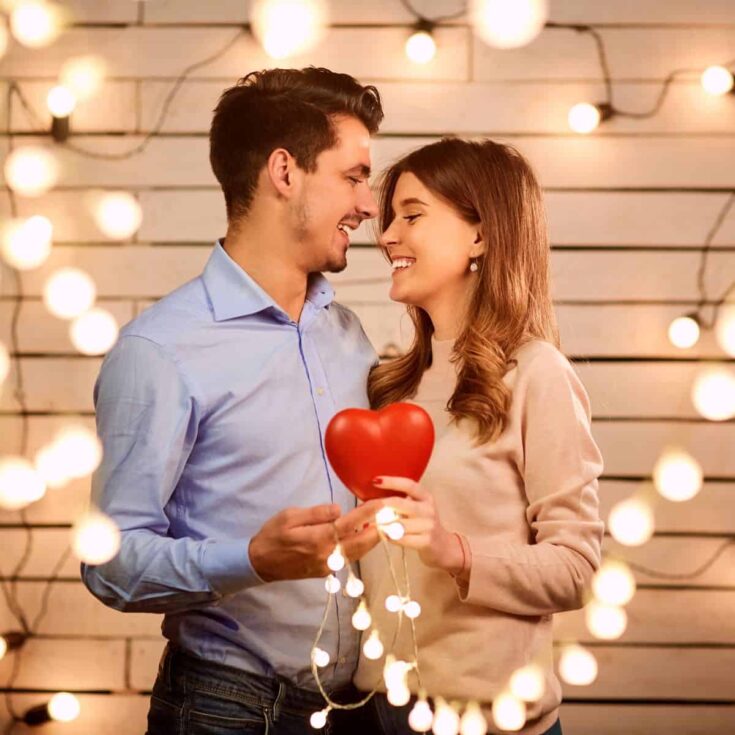 A couple smiling at each other while holding a red heart as they are being surrounded by fairy lights: date jar ideas