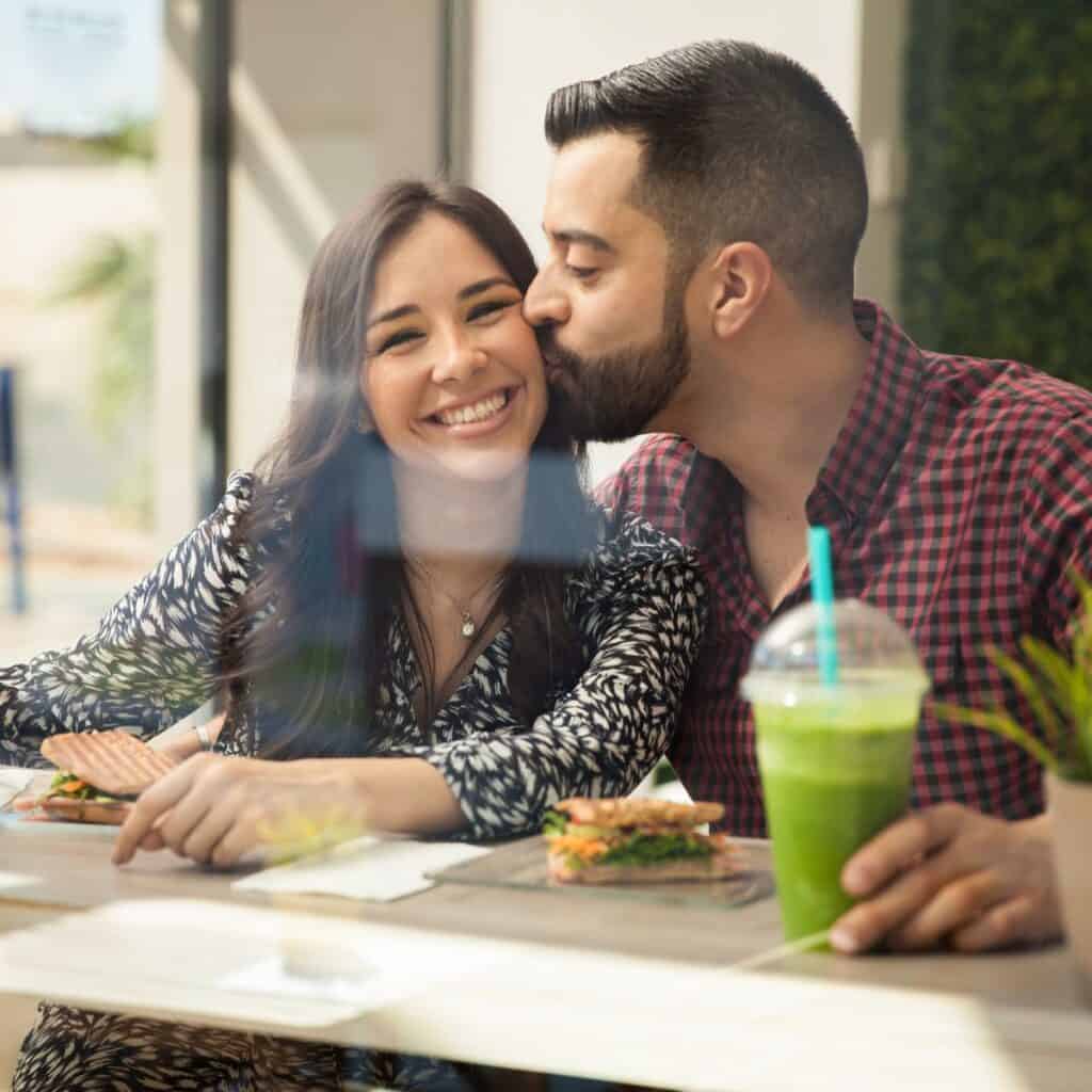 A man kissing on the woman's cheeks while they have lunch together after reading these lunch date ideas for busy couples