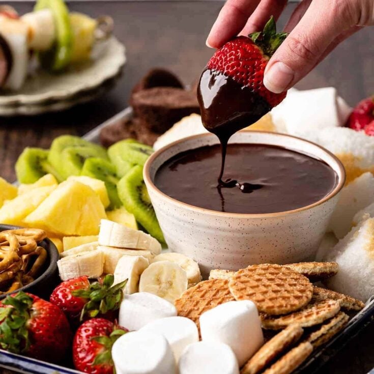 A stawberry being dipped on a chocolate fondue bowl, surrounded by fruits, biscuits, marshmallows, and more, made after reading these Easy fondue recipes