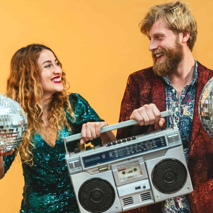 A happy couple wearing sparkly clothes, holding a big radio and mirror balls, after reading these 70s date night ideas