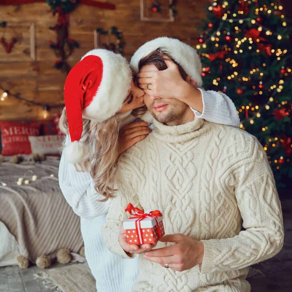 A woman kissing the man's cheeks while covering his eyes, the couple making a new tradition after reading these Christmas date ideas