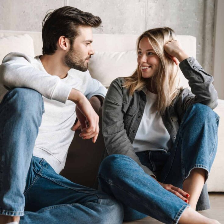 A couple sitting on the floor of their living room, asking each other these hypothetical questions for couples