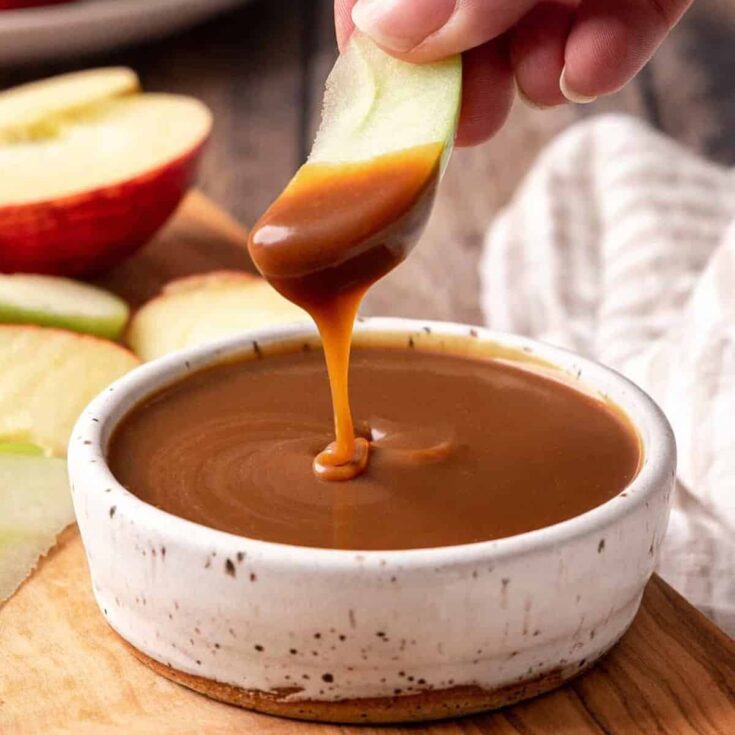 A small bowl of caramel apple dip, with a person dipping an apple to it