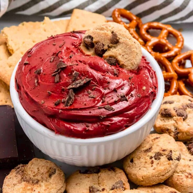 A bowl of red velvet cheesecake dip sided with cookies, biscuits, and pretzels