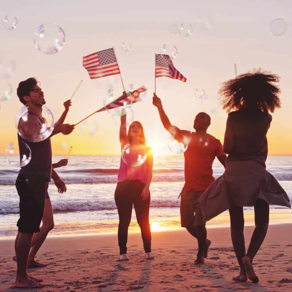 A group of friends at a beach, dancing around while each holding an American flag