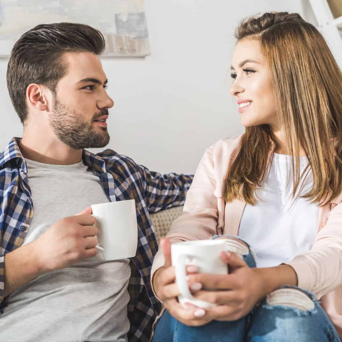 A happy couple sitting on the couch, both holding a cup of coffee, while asking each other these would you rather questions in-law edition